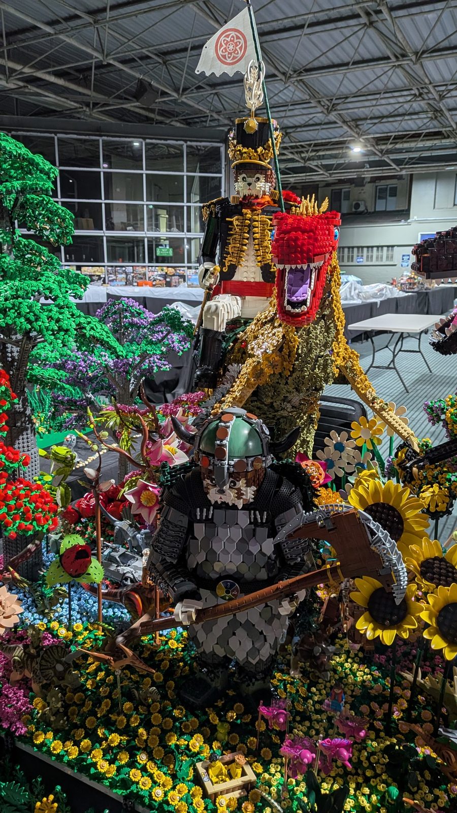 Large brick-built warrior figures and a red-headed dinosaur among LEGO flowers and trees